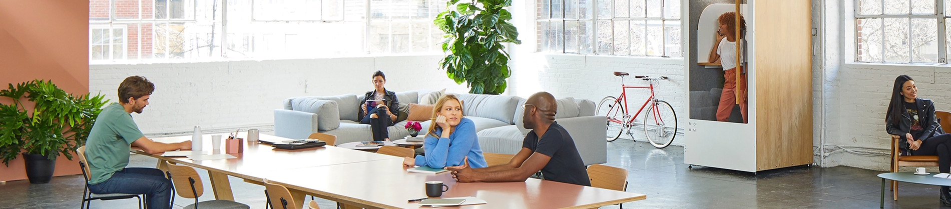 Two work colleagues talking at a table with other colleagues sat around the office Two work colleagues talking at a table with other colleagues sat around the office