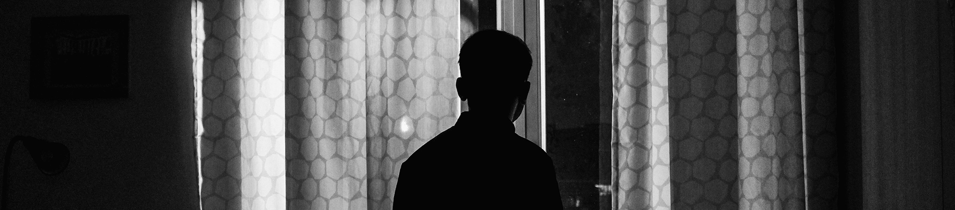A black and white photo of a man looking out of a window with the curtains slightly open A black and white photo of a man looking out of a window with the curtains slightly open