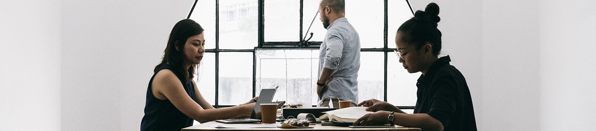 Two women sat at a table looking over paperwork with a guy stood looking out of the window behind them