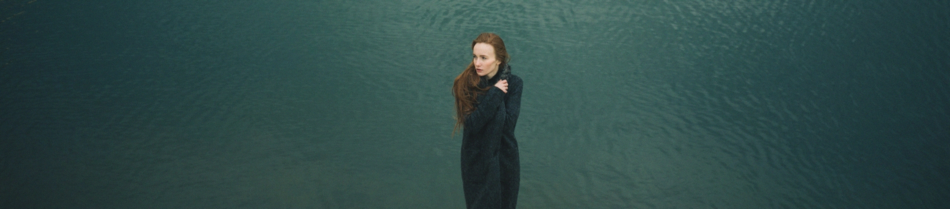 A women stood huddled in her cardigan with the ocean behind her