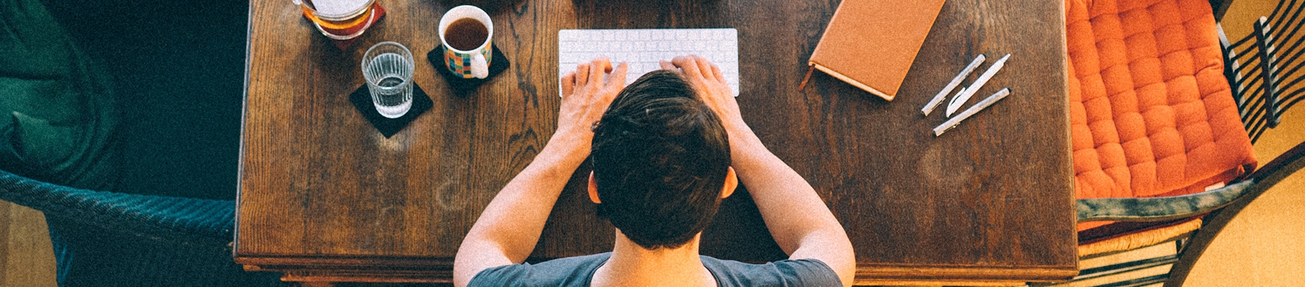 A guy sat at a wooden table typing on a keyboard with a notebook, pens and drinks A guy sat at a wooden table typing on a keyboard with a notebook, pens and drinks