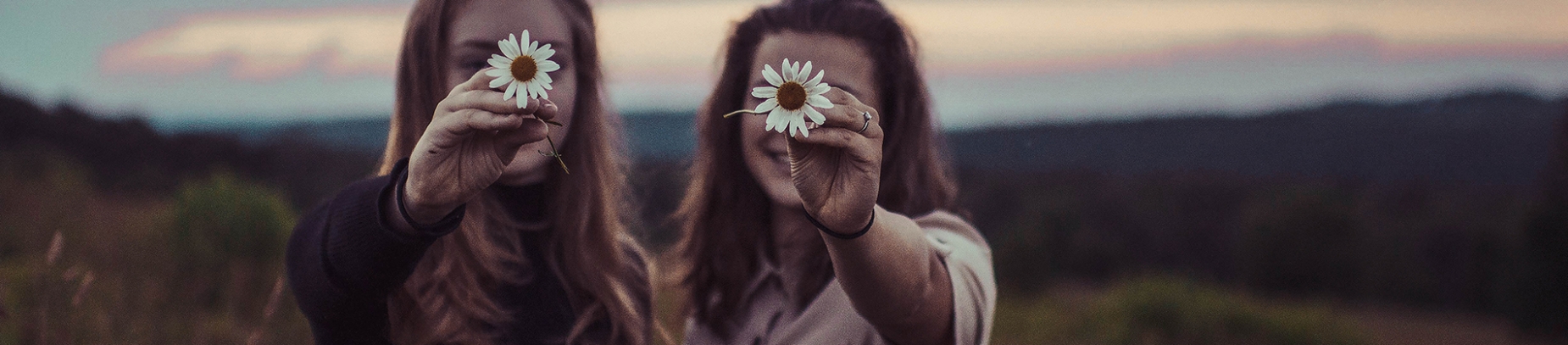 Two girls stood in a field holding up flowers in front of their faces Two girls stood in a field holding up flowers in front of their faces