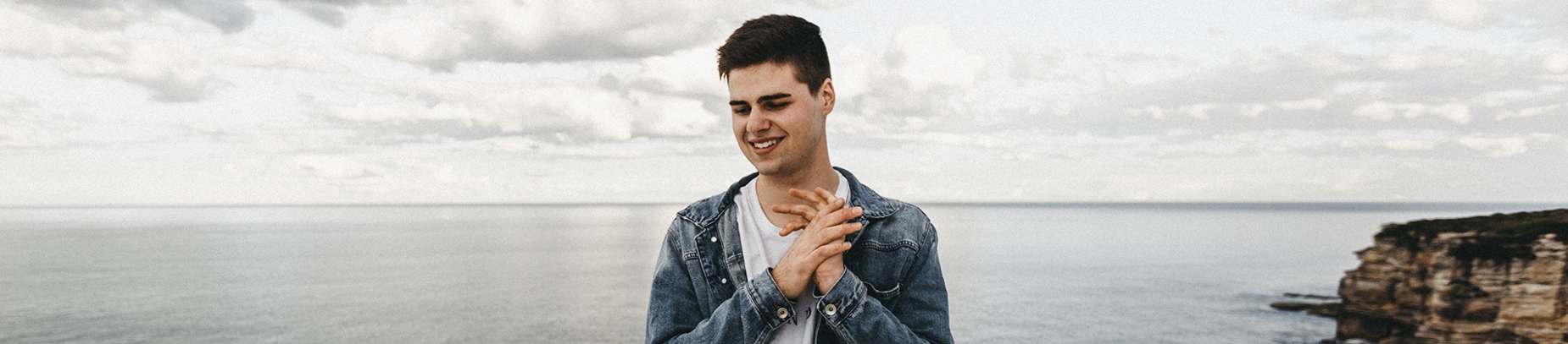 A guy smiling with his hands clasped with the ocean behind him