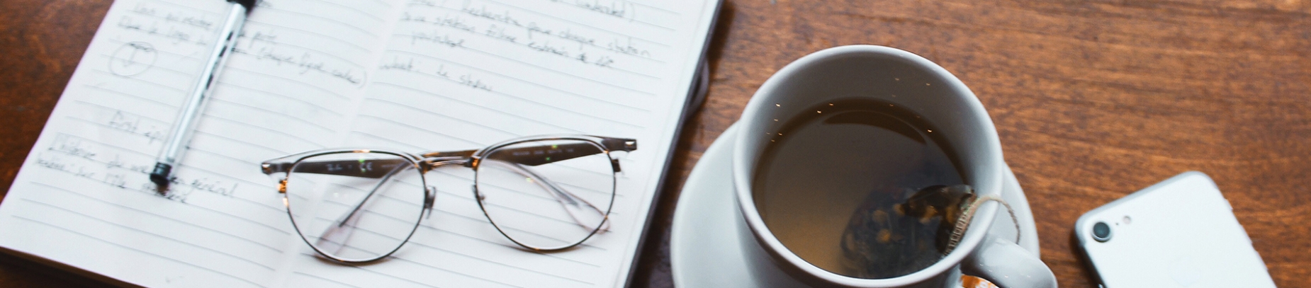 An open notebook and pen on a wooden table with a cup of tea, phone and glasses