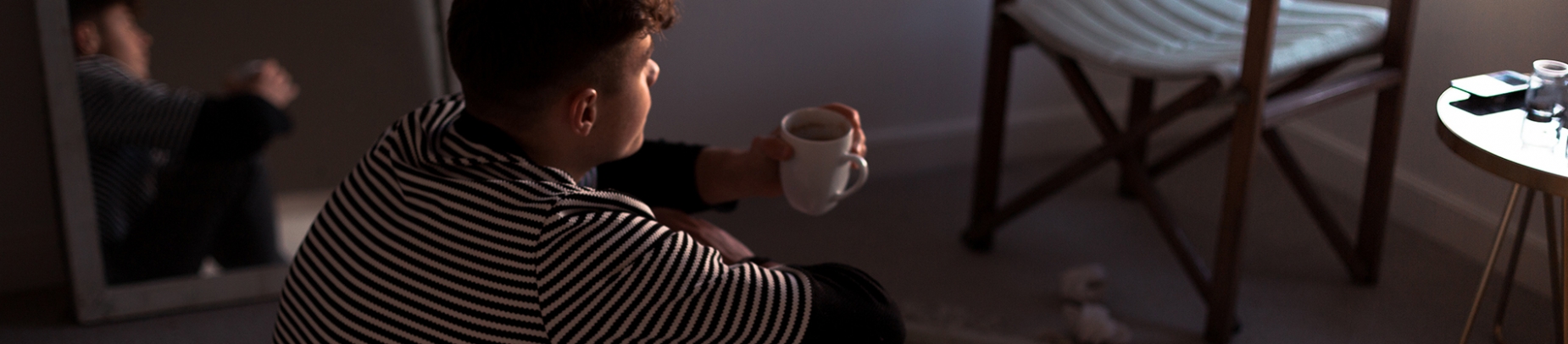 A young man sat on his bedroom floor with a cup of coffee