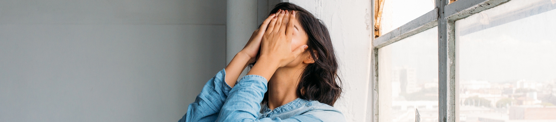 A women in a blue top with her hands over her face against a window